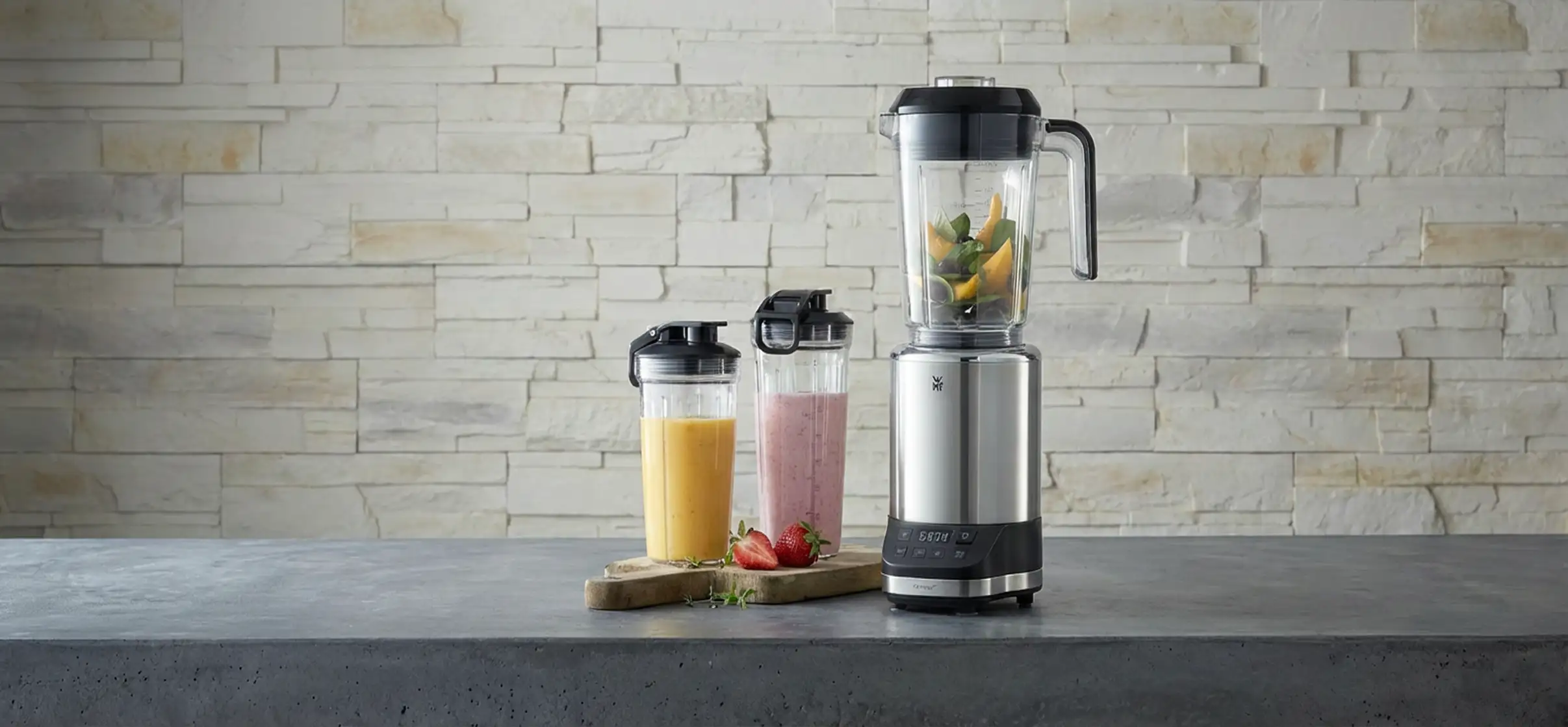 Mixer in silber gefüllt mit Obst und daneben zwei Shaker-Flaschen gefüllt mit Saft