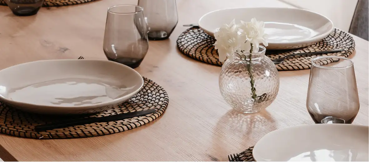 Gedeckter Tisch mit hellen Tellern und runden Tischsets. Daneben Gläser und eine Glas-Vase mit Blume