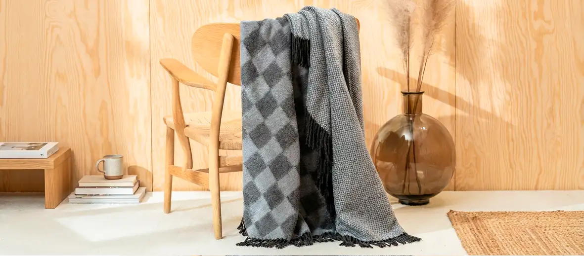 Dünne Decke in grau gemustert über Holzstuhl hängend. Vase in braun mit Pampasgras