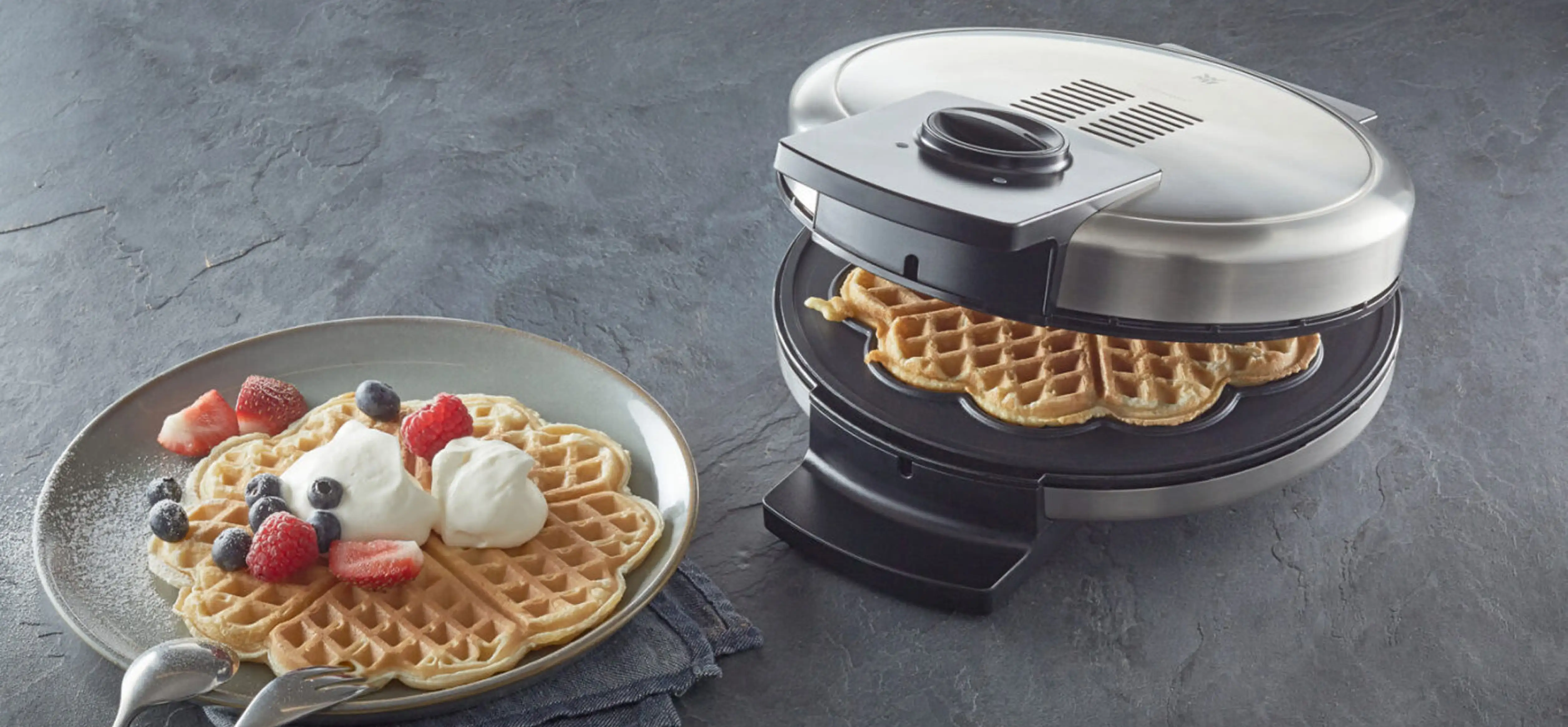 Waffeleisen in silber mit einer gebackenen Waffel. Daneben ein Teller mit angerichteter Waffel mit Sahne und Beeren