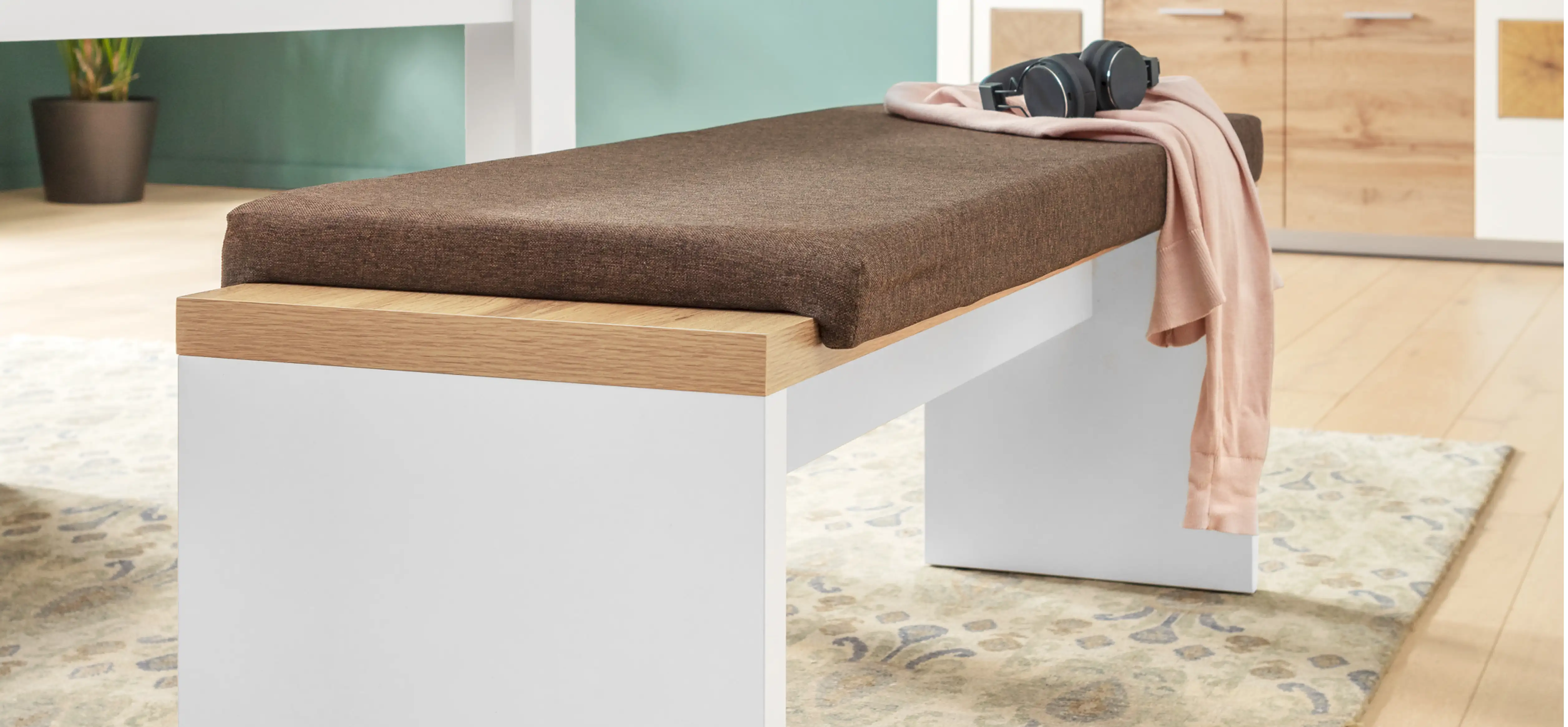 Sitzbank aus weißem Holz und naturfarbenen Sitzfläche. Darauf ein Sitzkissen in dunklem Braun