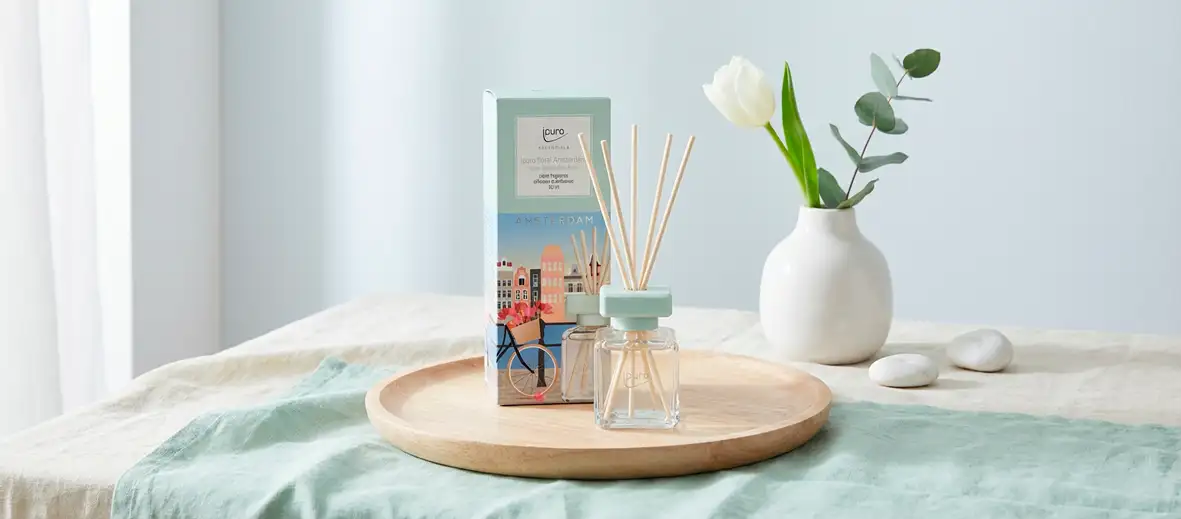 Raumduft in türkis auf Holztablett, dahinter eine Vase mit Blumen