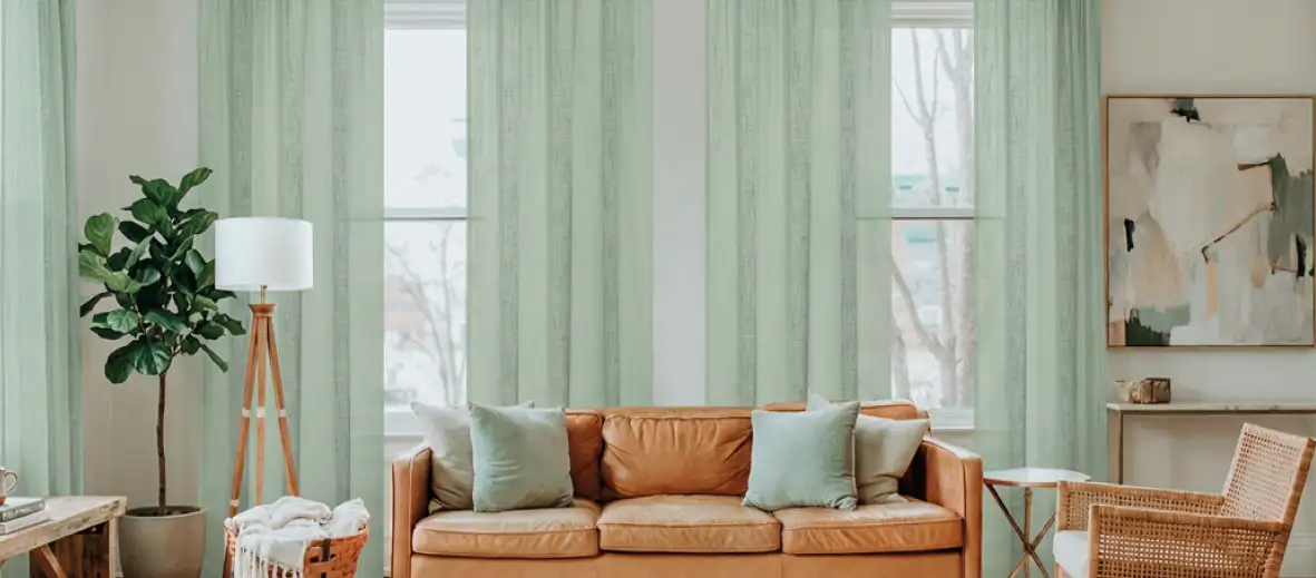 Vorhänge in Minze vor großen Fenstern. Davor Ledersofa in braun mit passenden Kissen. Stehlampe, Pflanze und Sessel daneben