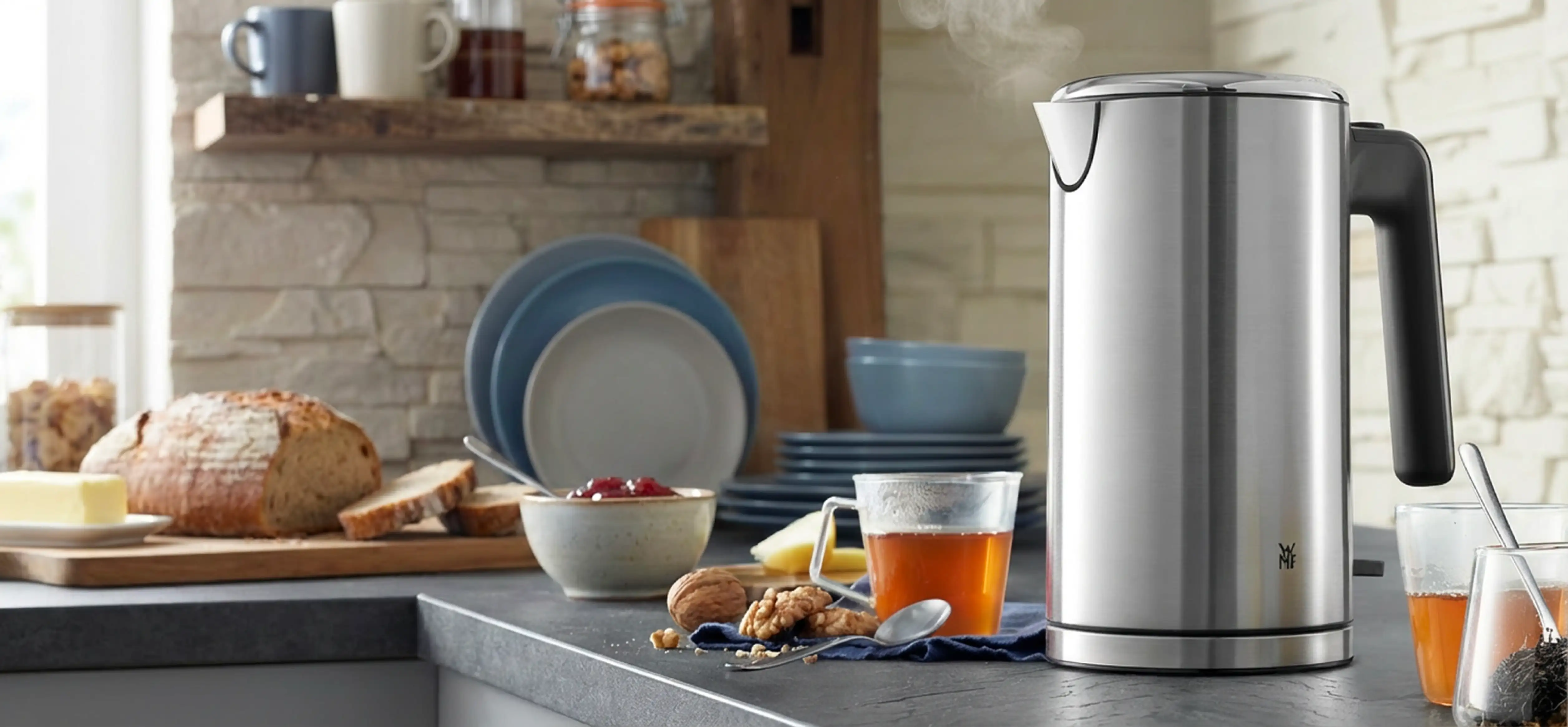 Wasserkocher in silber mit gefülltem Teeglas daneben. Im Hintergrund Brot, Lebensmittel und Geschirr